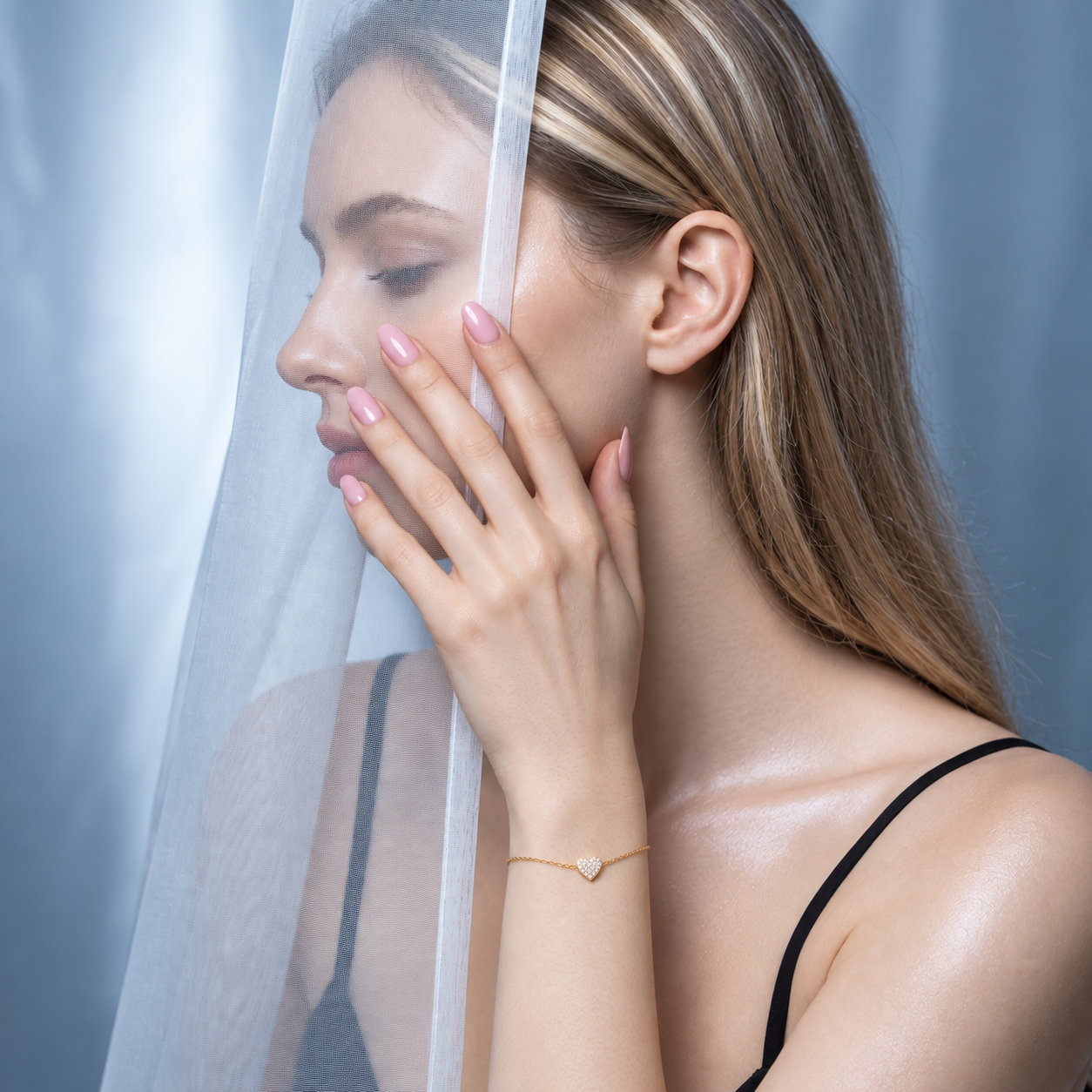A woman in a black camisole wears a delicate gold chain bracelet with a small heart-shaped pearl pendant, posed under a sheer white curtain.