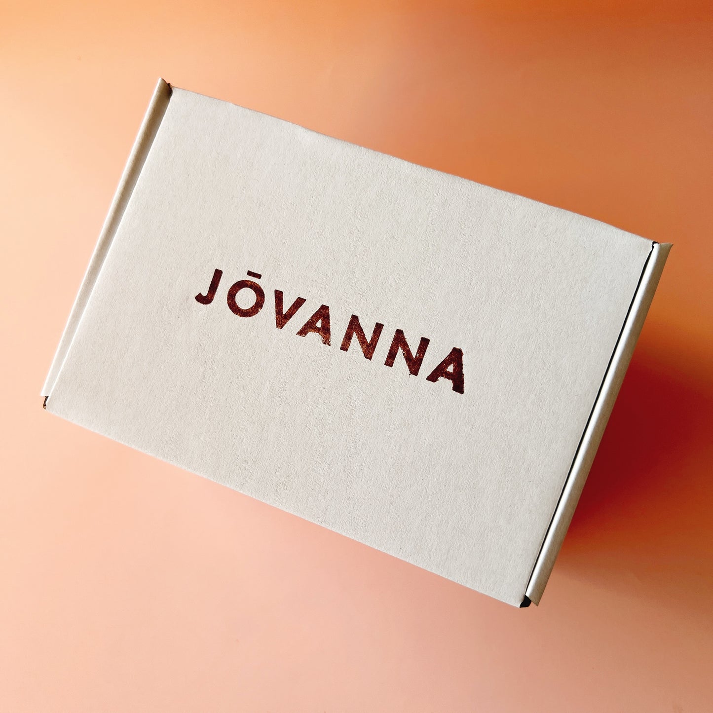 A white cardboard box with the brand name 'JÓVANNA' printed in dark red across the top, photographed at an angle against a warm peach-orange gradient background.