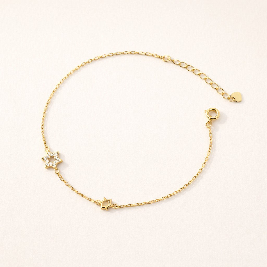 A delicate gold chain bracelet with two small star charms, one adorned with a tiny clear crystal, displayed on a light beige textured background.