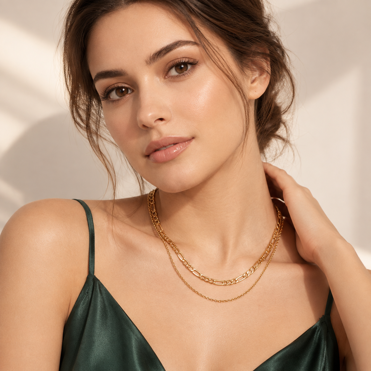 A close-up lifestyle shot of a woman wearing a double-layered gold chain necklace, including a chunky curb link chain and a delicate fine link chain. She is wearing a dark green satin camisole top against a warm, softly lit beige background.