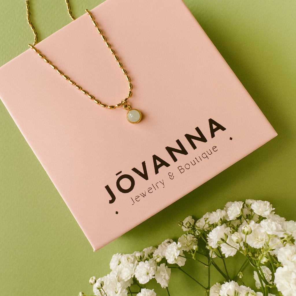 Celadon Bloom Necklace draped over a linen box with white flowers, mint-green pendant in gold bezel