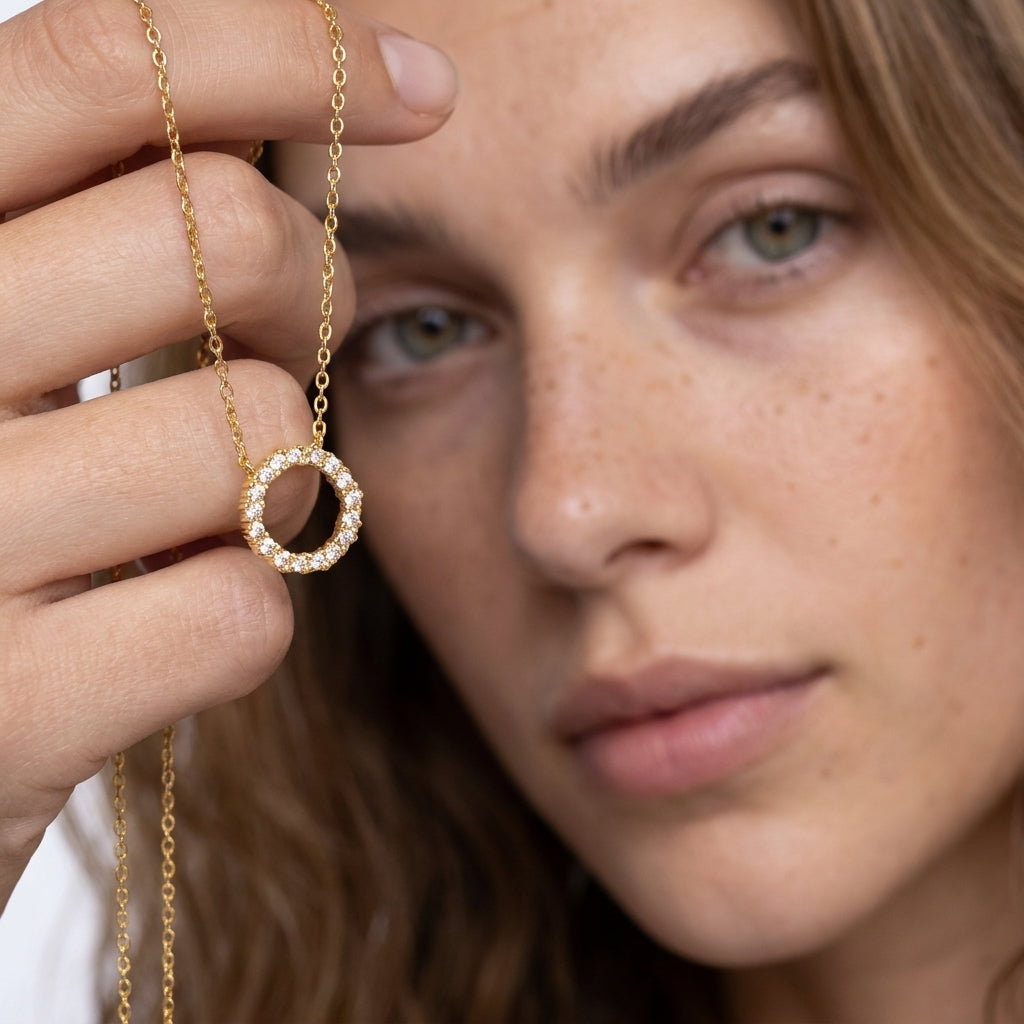 A close-up lifestyle shot of a woman holding a delicate gold chain necklace with a circular ring pendant adorned with small clear stones.