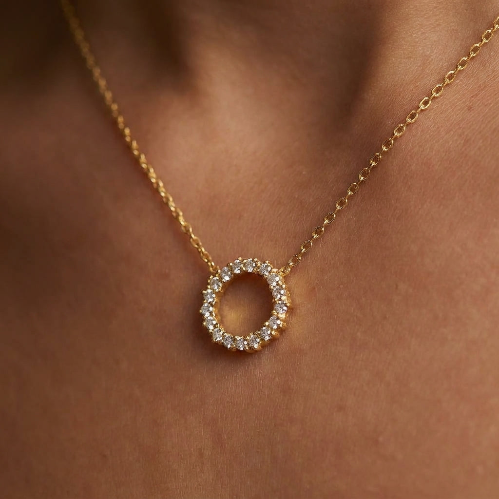 A delicate gold chain necklace with an open circular pendant encrusted with small clear stones, shown worn on a person's neck against a warm skin-toned background.