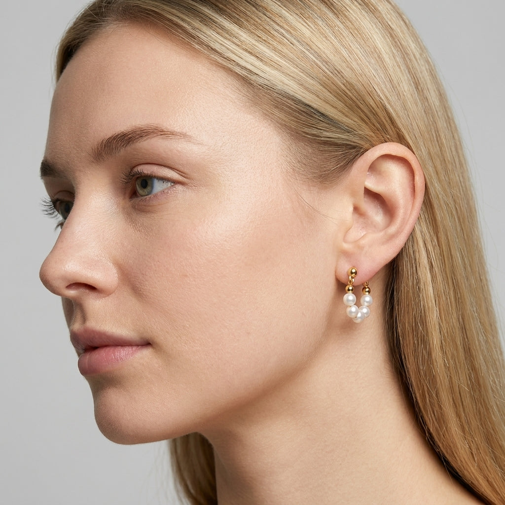 A close-up side profile of a blonde woman wearing a delicate gold hoop earring with a small cluster of white pearls hanging from it.