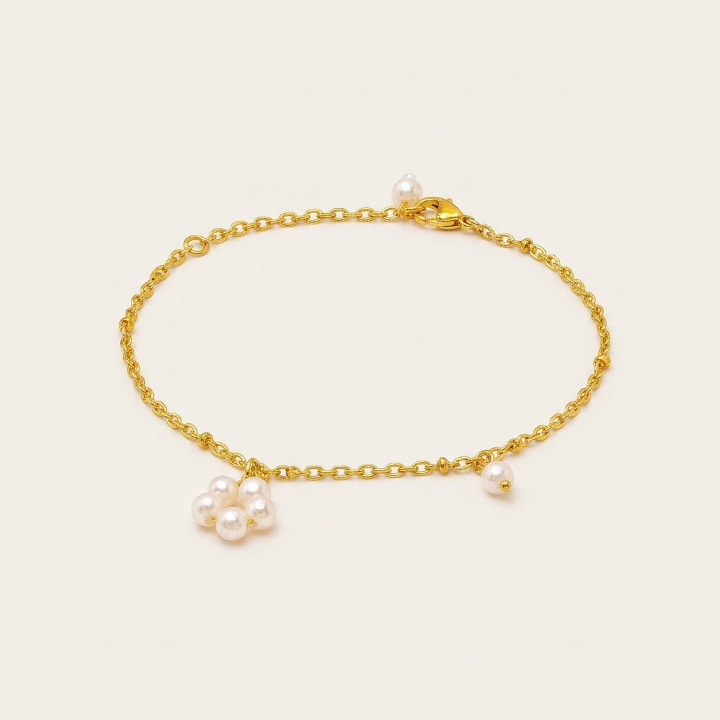 A delicate gold chain bracelet featuring a small cluster of white freshwater pearls and a single loose pearl charm, displayed on a light cream background.
