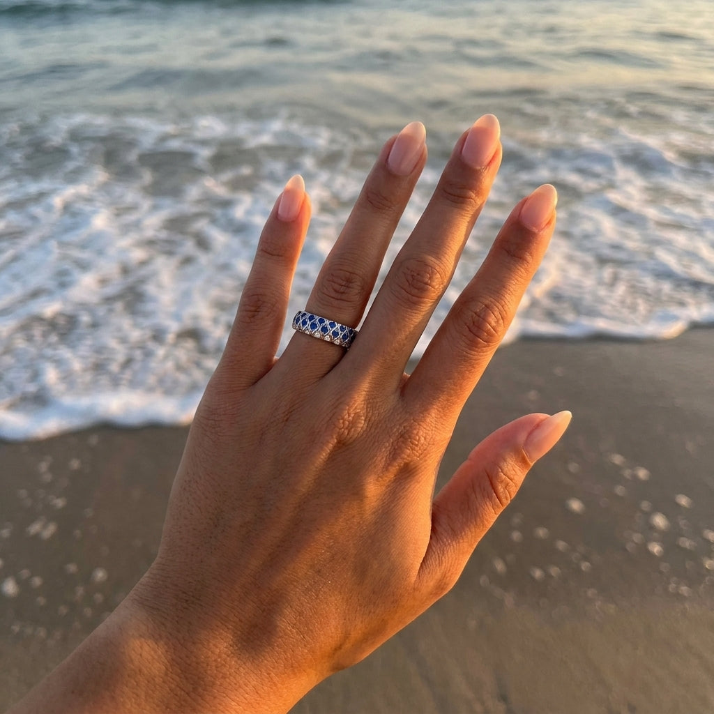 Hand wearing the Azure Sea Ring on the middle finger, sterling silver eternity band with blue cubic zirconia, ocean waves in the background