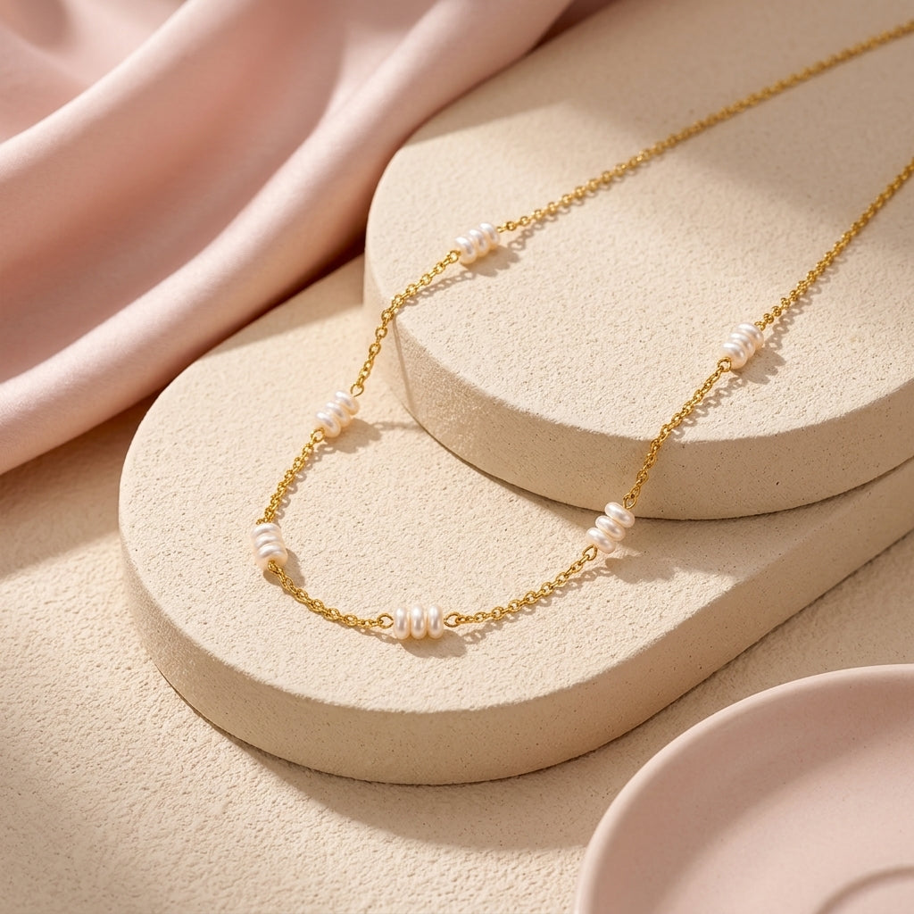 Pearl Moments Necklace, a gold chain with groups of three small white pearls, laid on cream stone stands with soft pink fabric
