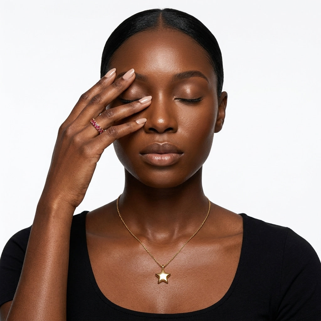 Model wearing the gold Little Star Pendant Necklace at the collarbone with a black scoop-neck top, eyes closed and hand resting on her face. 