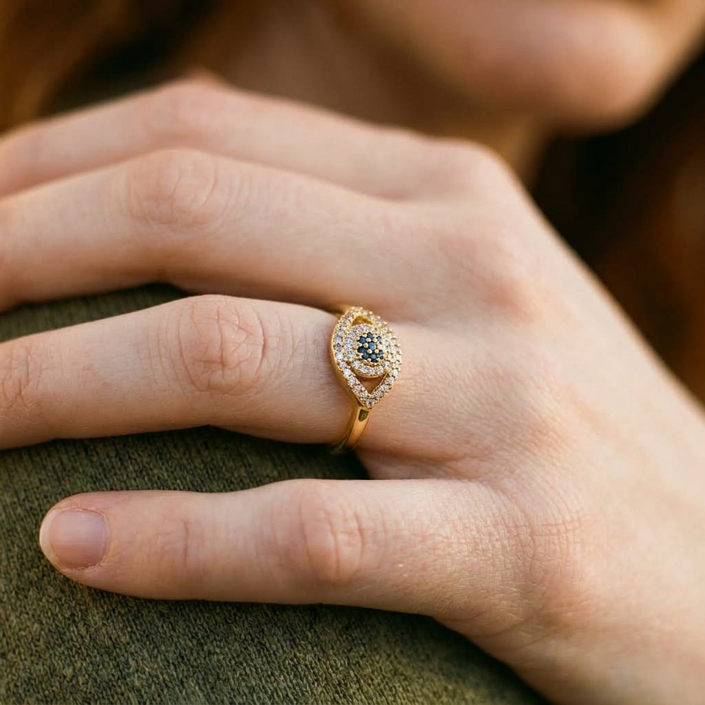 A gold-toned eye-shaped ring with a central blue gemstone and small clear stones, worn on a person's finger.